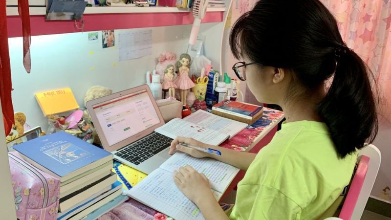 Hình ảnh một bạn nam đang chăm chỉ học trên laptop và ghi chép, với nhiều sách xung quanh, thể hiện sự miệt mài trong học tập online hoặc nghiên cứu